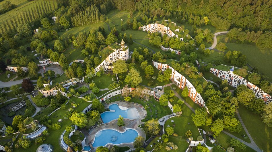 Vogelperspektive auf das Rogner Bad Blumau im Thermen- & Vulkanland Steiermark mit Hundertwasser-Architektur, Thermalbecken, Hotelanlage, Parklandschaft und umgebendem Wald als Wellnessdestination.