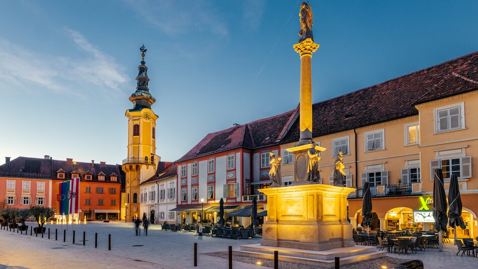 Hauptplatz von Bad Radkersburg in der Südoststeiermark mit beleuchteter Mariensäule, historischem Stadtturm, Altstadtfassaden und Gastronomie am Abend.