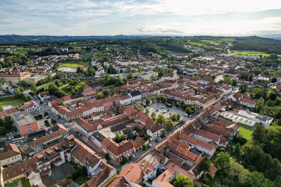 Luftaufnahme der historischen Altstadt von Fürstenfeld im Thermen- & Vulkanland Steiermark mit geschlossenem Häuserensemble, rotdachigen Bürgerhäusern, Hauptplatz und umliegender grüner Hügellandschaft an einem sonnigen Tag.