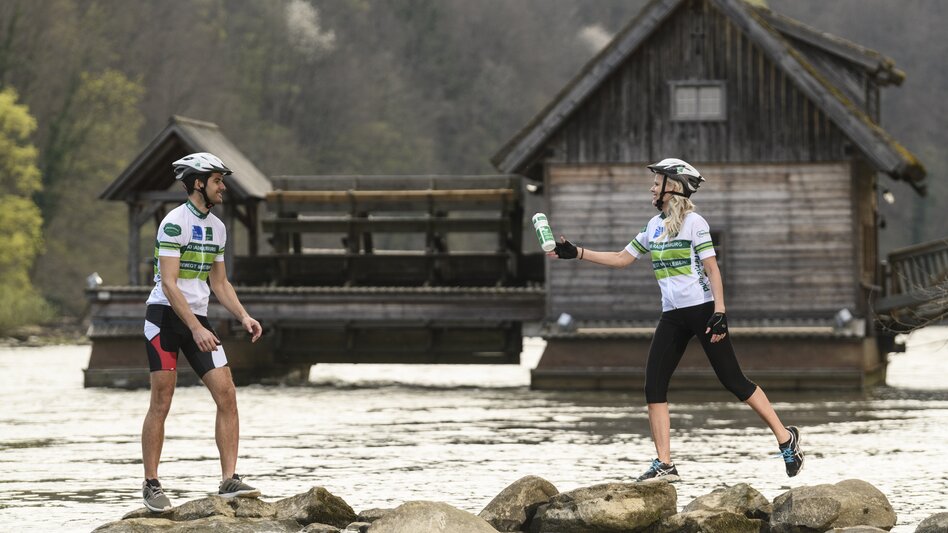 Radfahrer werfen Sportflasche in die Luft, Schiffsmühle Mureck im Hintergrund | © TVB Thermen- & Vulkanland | pixelmaker.at