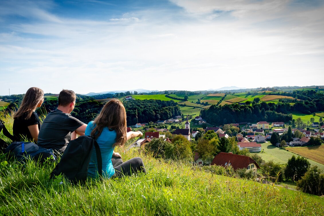 Zwei Frauen und ein Mann sitzen auf der grünen, saftigen Wiese und blicken auf Unterlamm im Thermen- & Vulkanland Steiermark