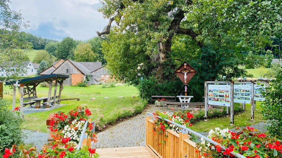 Blumengeschmückter Rastplatz mit Holzbrücke, Sitzgelegenheiten und altem Baumbestand in Unterlamm im Thermen- & Vulkanland.