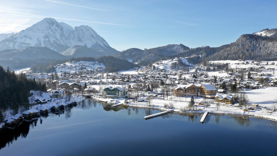 Winterliche Luftaufnahme von Altaussee: Der tiefblaue See liegt ruhig zwischen schneebedeckten Bergen, umgeben von verschneiten Wäldern und dem kleinen Ort mit seinen Häusern am Ufer. | © Tom Lamm