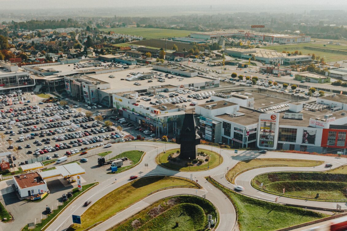Luftaufnahme der ShoppingCity Seiersberg bei Graz mit großen Parkplätzen, umliegenden Straßen und angrenzender Autobahn im Vordergrund.