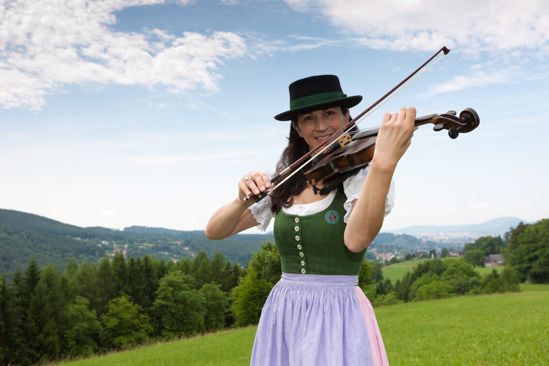 Frau in Tracht spielt Geige auf Wiese bei Stattegg, Hügel und Landschaft der Steiermark im Hintergrund