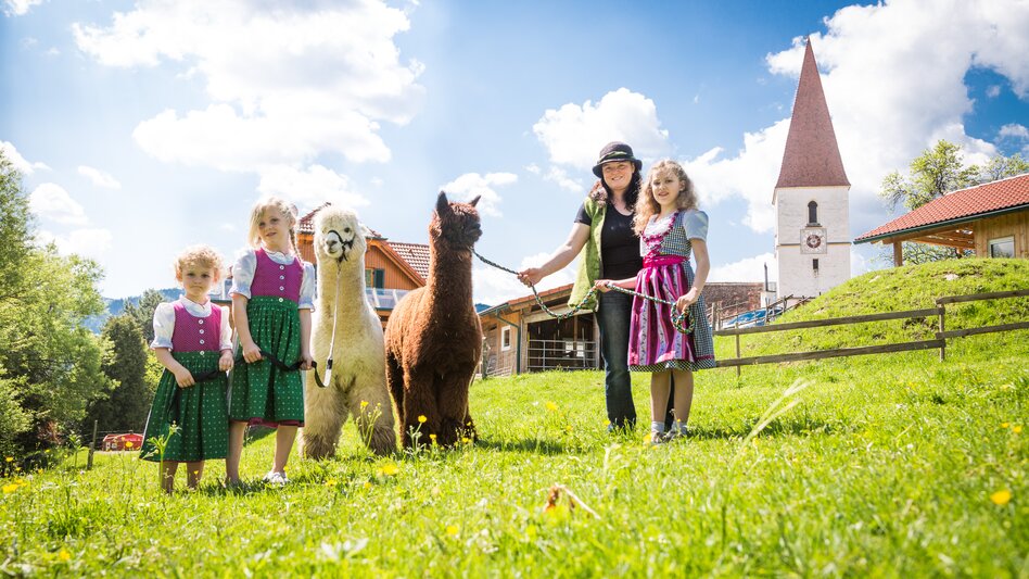 Eine Frau und einige Kinder stehen mit flauschigen Alpakas auf einer blühenden Sommerwiese