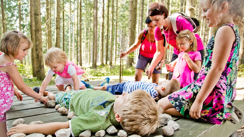 Zwei Kinder liegen auf einer Holzliege im Wald, Kinder legen Steine rundherum