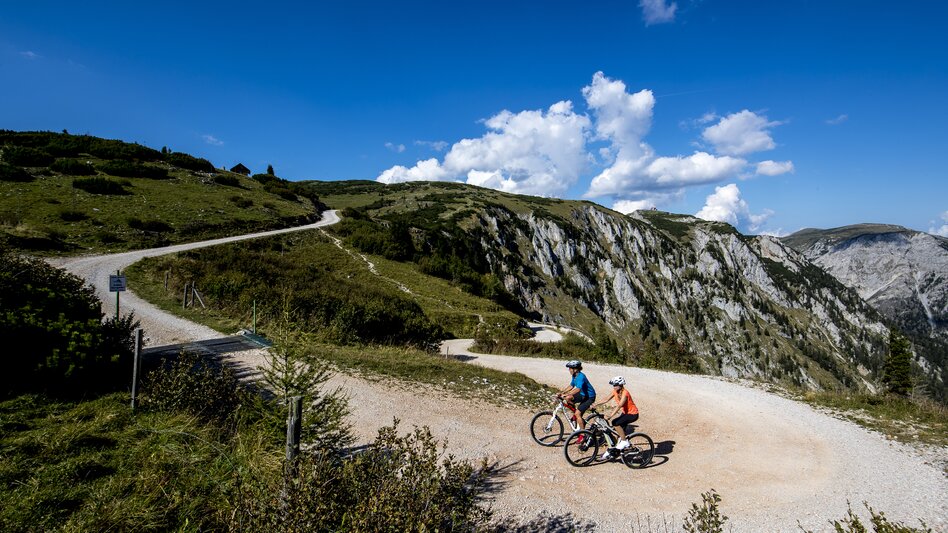 Umrahmt von einer Bergkulisse fahren zwei Radfahrer die Schotterstraße hinauf.