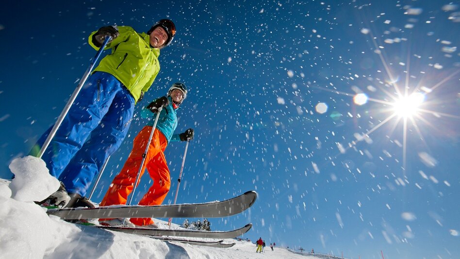 Zwei Skifahrer stehen an der Piste, im Fokus die Skier, der Schnee staubt und sie lachen