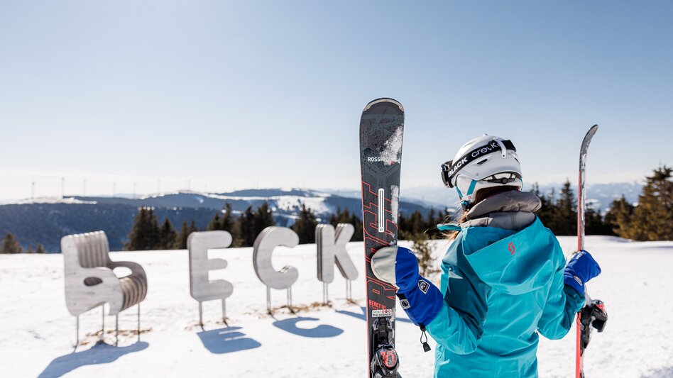 Eine Frau hält ihre beiden Skier in den Händen, im Hintergrund der Schriftzug des Skigebiets aus Eisen im Schnee