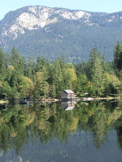 Ein ruhiger See umgeben von dichtem Wald und Bergen. Die Hütten spiegeln sich im klaren Wasser. | © TVB Ausseerland Salzkammergut-Tom Lamm
