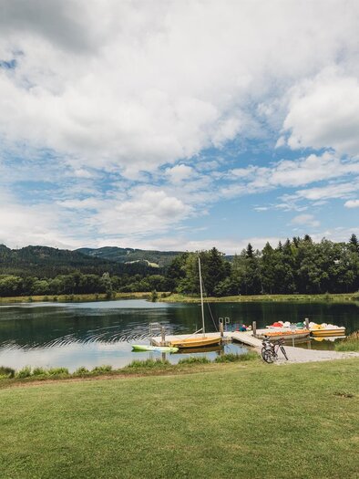 A calm lake with boats and green grass. In the background are gentle hills and a beautiful sky. | © nicoleseiser.at