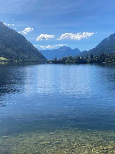 Der ruhige Grundlsee aus Richtung Gößl fotografiert, umgeben von den Bergen des Ausseerland Salzkammergutes. Der Himmel ist klar und blau, und das Wasser spiegelt die wunderschöne Landschaft wider. | © Bettina Scheck