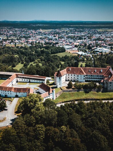 An impressive view of a historic castle, surrounded by green forests and a city in the background. The architecture is distinctive and well-preserved. | © Patrick Schadler | shutterstock.com