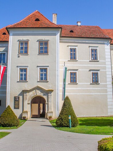 Ein historisches Gebäude mit roten Dächern und großen Fenstern. Der Eingang ist von gepflegten Bäumen und Sträuchern umgeben. | © Sandra Brünner